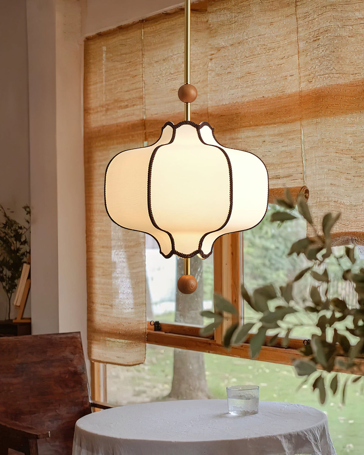 Lit white lantern pendant above a cloth-covered table by a timber window onto a garden.