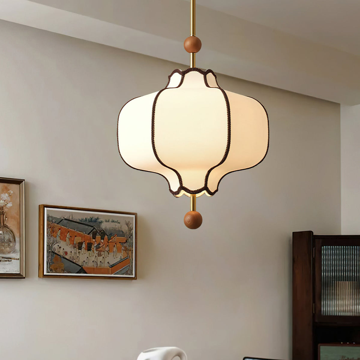 Lit white fabric lantern pendant with brown braid trim hanging beside two framed paintings.