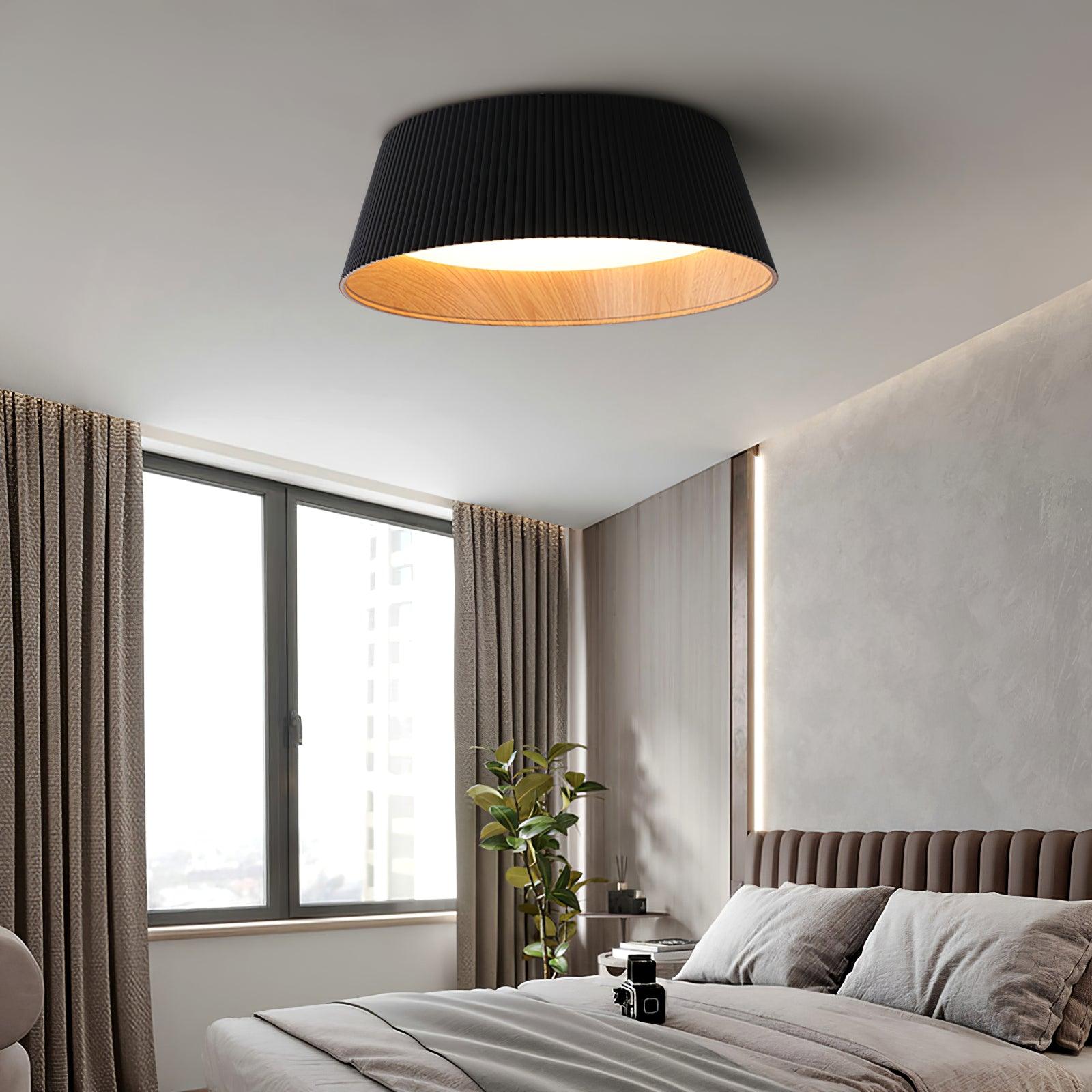 Drum-shaped flush ceiling light with a ribbed black outer, a wood-look inner rim and a white diffuser fitted above a bed by a window.