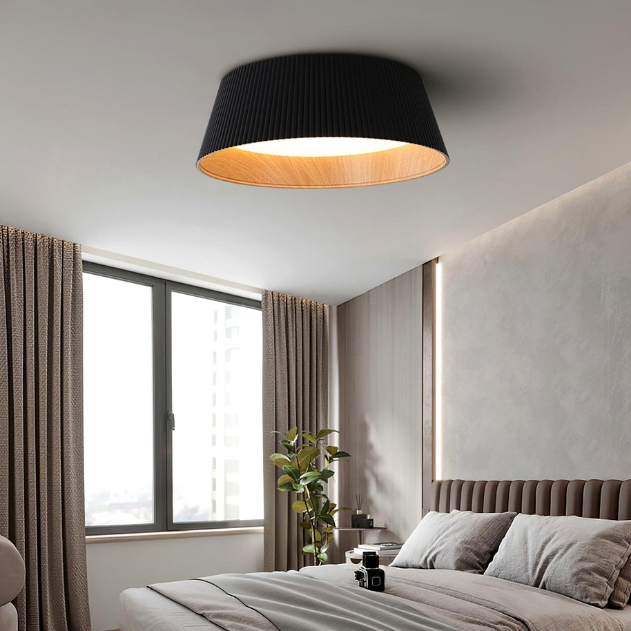 Drum-shaped flush ceiling light with a ribbed black outer, a wood-look inner rim and a white diffuser fitted above a bed by a window.