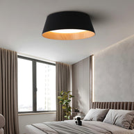 Drum-shaped flush ceiling light with a ribbed black outer, a wood-look inner rim and a white diffuser fitted above a bed by a window.