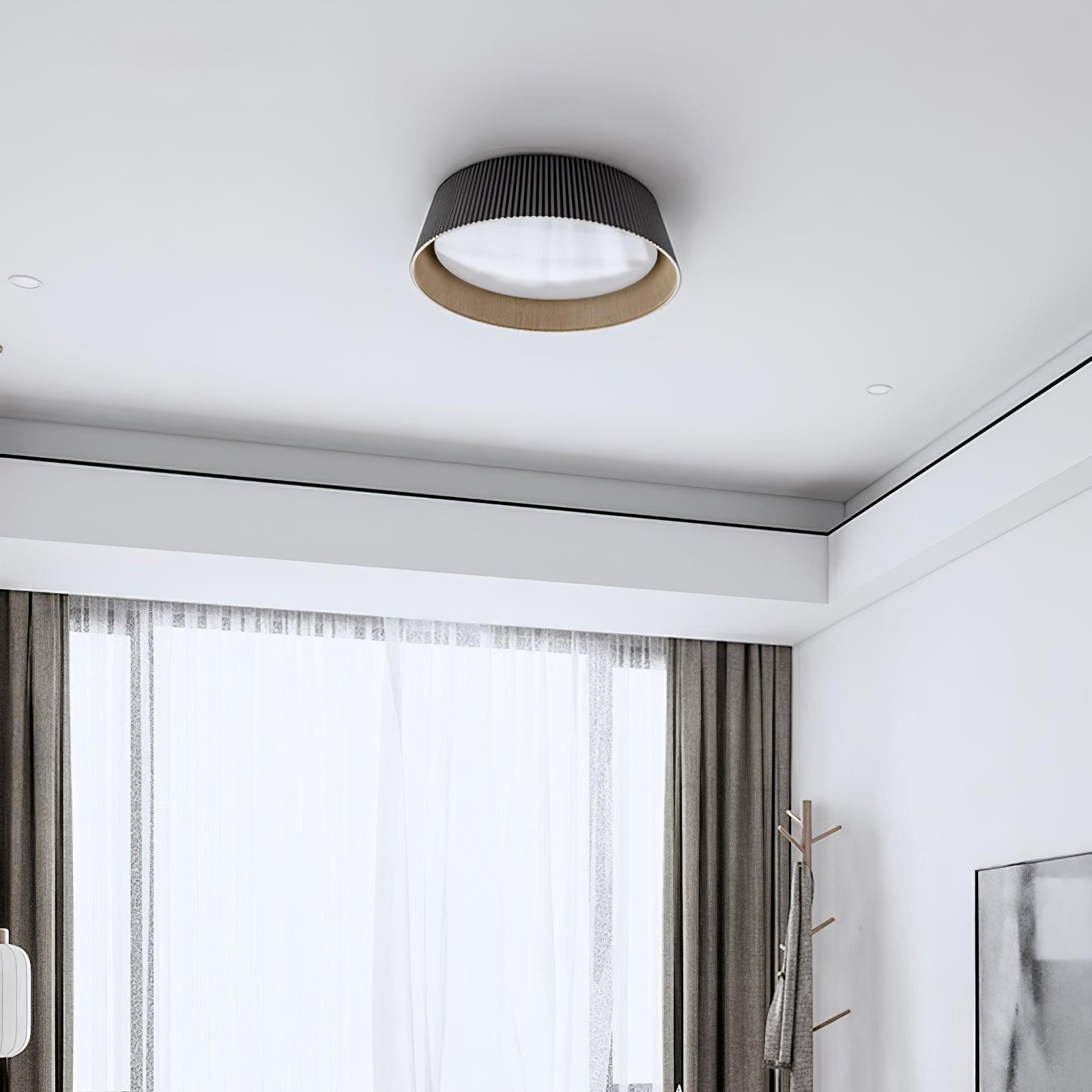 Drum-shaped flush ceiling light with a ribbed black outer, a wood-look inner rim and a white diffuser fitted to a ceiling beside a curtained window.