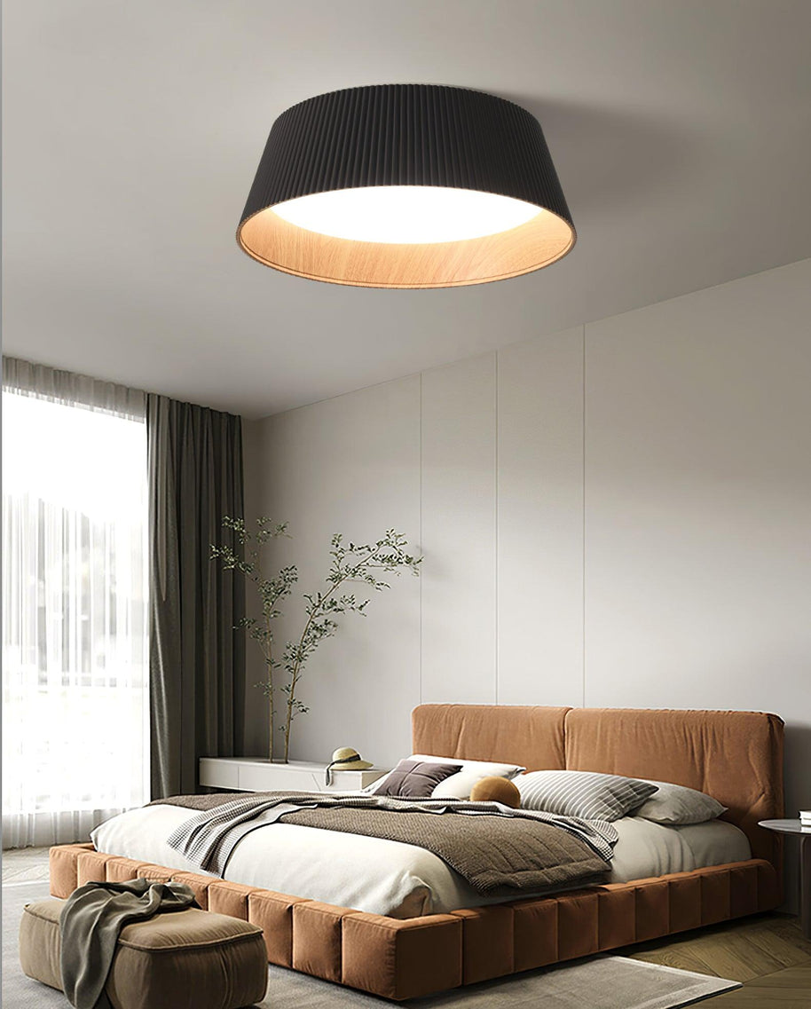 Drum-shaped flush ceiling light with a ribbed black outer, a wood-look inner rim and a white diffuser fitted above a bed in a room with a large window.