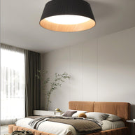 Drum-shaped flush ceiling light with a ribbed black outer, a wood-look inner rim and a white diffuser fitted above a bed in a room with a large window.