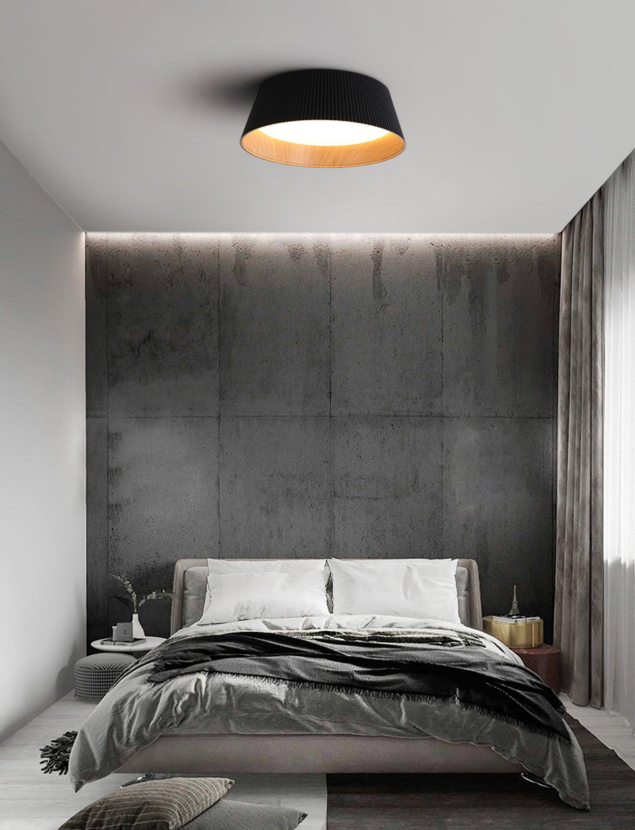 Drum-shaped flush ceiling light with a ribbed black outer, a wood-look inner rim and a white diffuser fitted above a bed against a textured wall.
