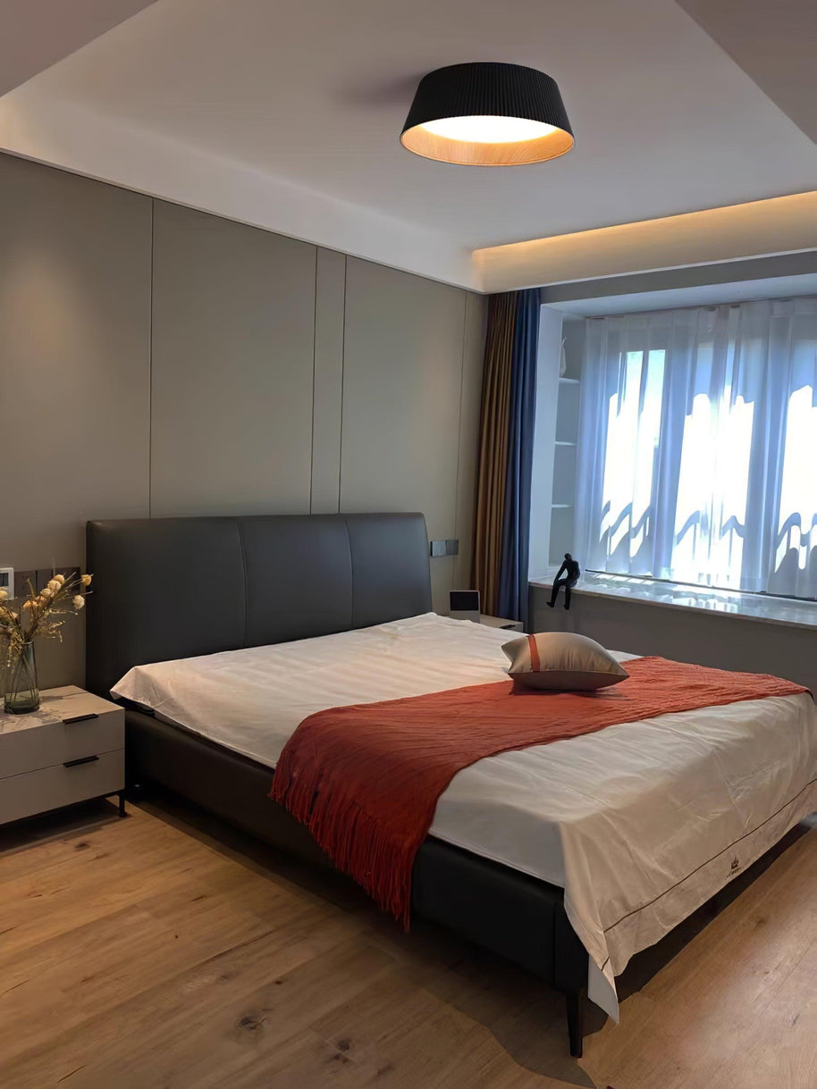 Drum-shaped flush ceiling light with a ribbed black outer, a wood-look inner rim and a white diffuser fitted above a bed with a red throw.