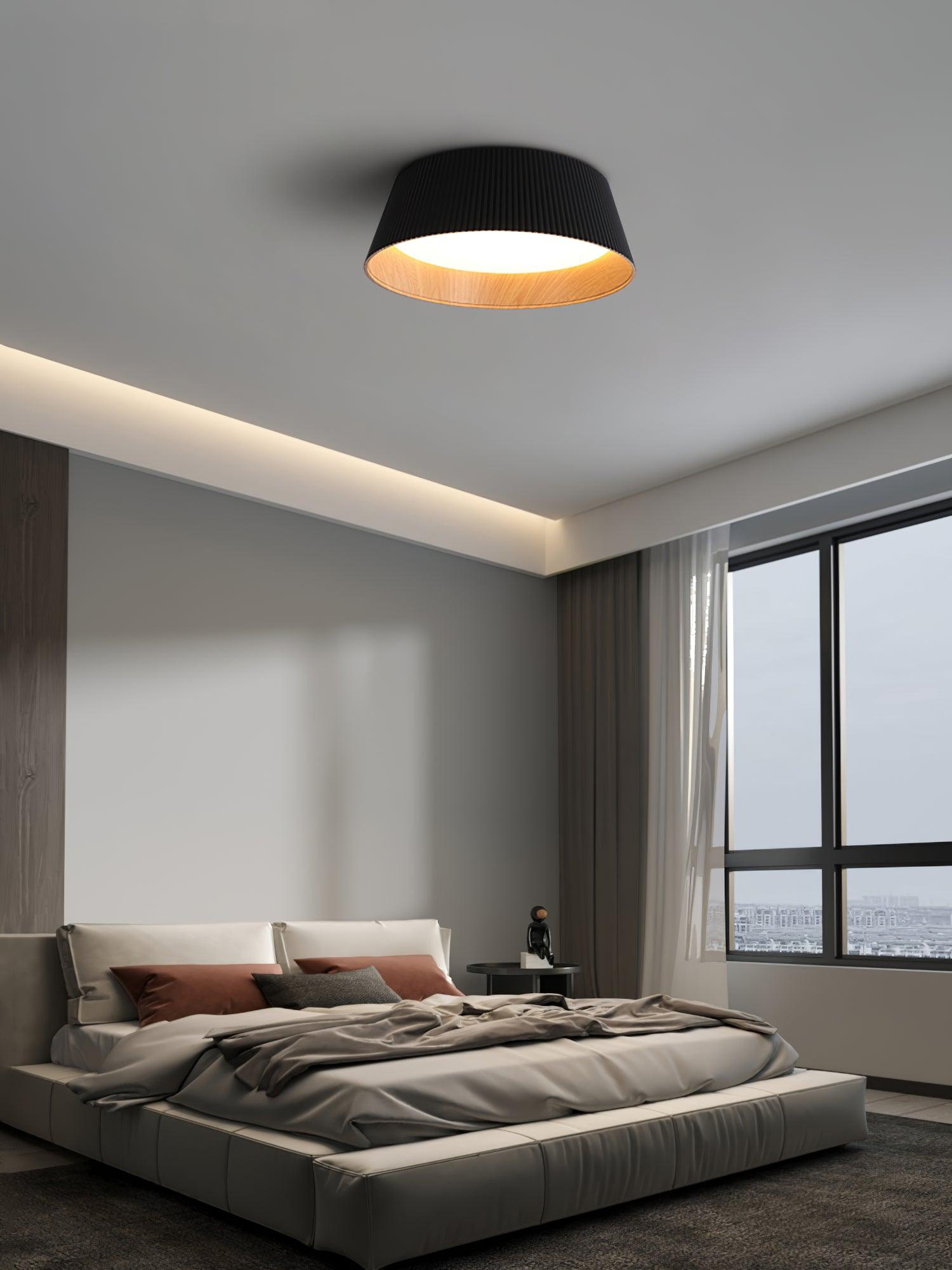 Drum-shaped flush ceiling light with a ribbed black outer, a wood-look inner rim and a white diffuser fitted above a bed by a window.