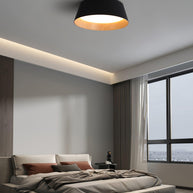 Drum-shaped flush ceiling light with a ribbed black outer, a wood-look inner rim and a white diffuser fitted above a bed by a window.