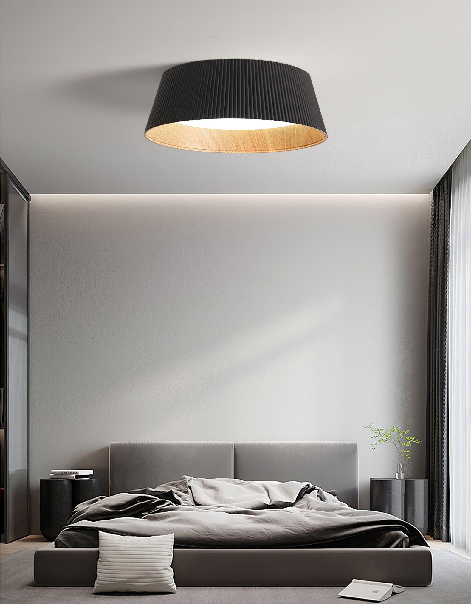 Drum-shaped flush ceiling light with a ribbed black outer, a wood-look inner rim and a white diffuser fitted above a bed against a panelled wall.