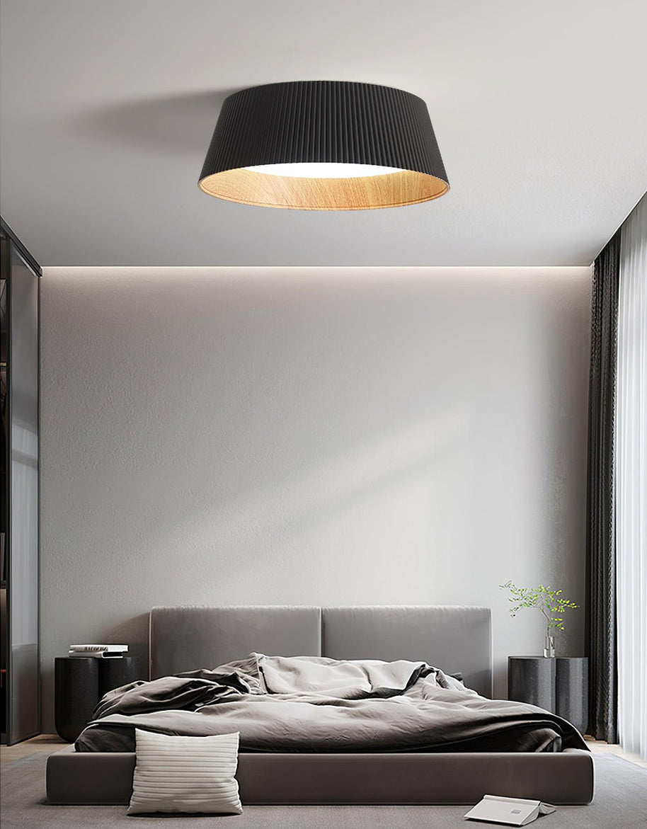 Drum-shaped flush ceiling light with a ribbed black outer, a wood-look inner rim and a white diffuser fitted above a bed against a panelled wall.
