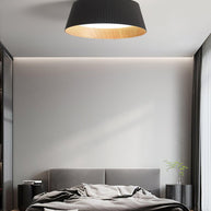 Drum-shaped flush ceiling light with a ribbed black outer, a wood-look inner rim and a white diffuser fitted above a bed against a panelled wall.