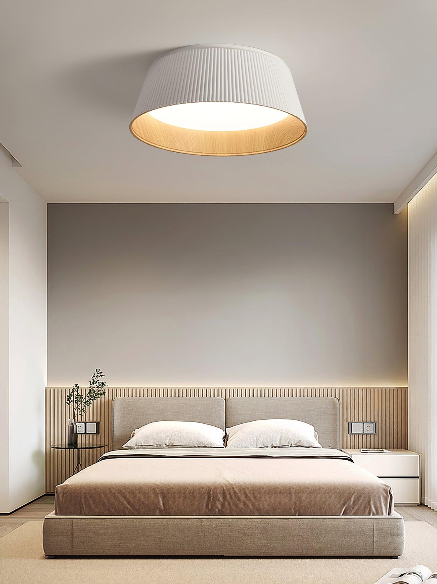 Drum-shaped flush ceiling light with a ribbed white outer, a wood-look inner rim and a white diffuser fitted above a bed with a slatted headboard.