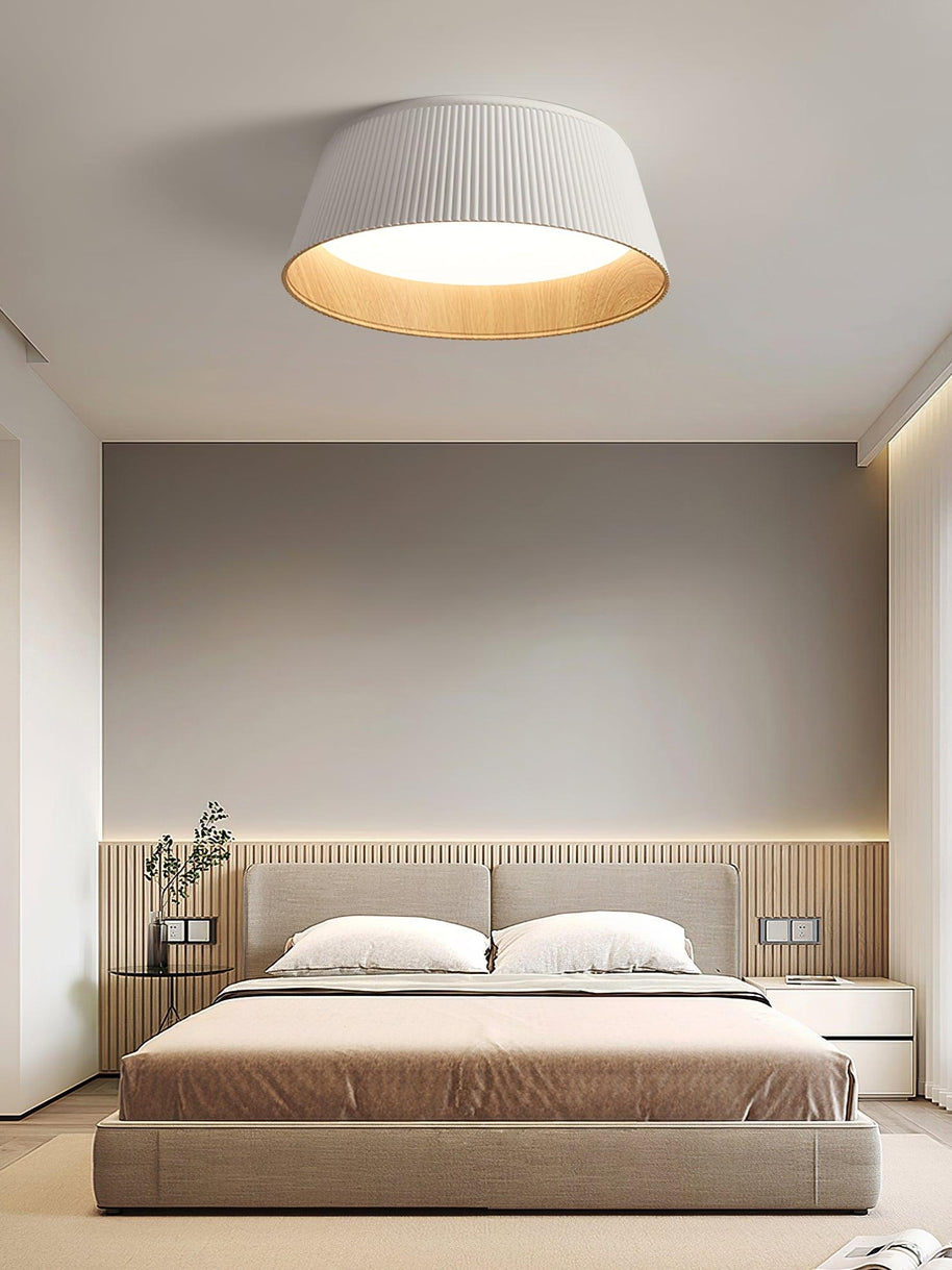 Drum-shaped flush ceiling light with a ribbed white outer, a wood-look inner rim and a white diffuser fitted above a bed with a slatted headboard.