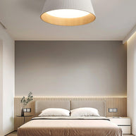 Drum-shaped flush ceiling light with a ribbed white outer, a wood-look inner rim and a white diffuser fitted above a bed with a slatted headboard.
