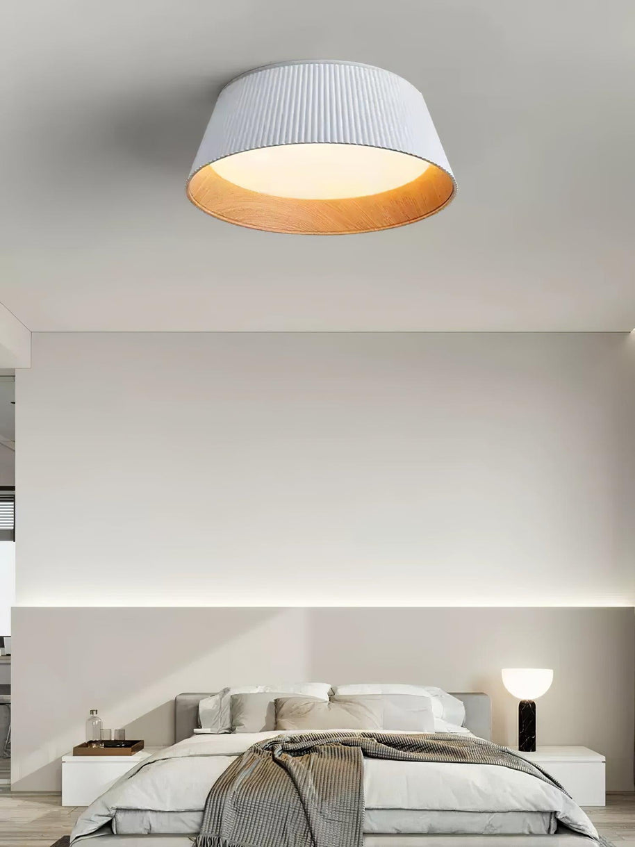 Drum-shaped flush ceiling light with a ribbed shade, a wood-look inner rim and a white diffuser, white ribbed shade, lit, fitted to a bedroom ceiling above a bed.