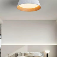 Drum-shaped flush ceiling light with a ribbed shade, a wood-look inner rim and a white diffuser, white ribbed shade, lit, fitted to a bedroom ceiling above a bed.