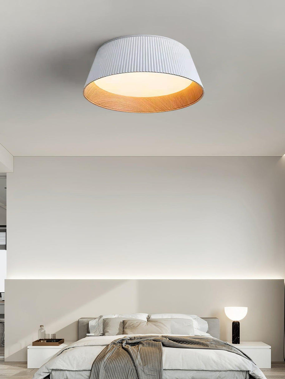 Drum-shaped flush ceiling light with a ribbed white outer, a wood-look inner rim and a white diffuser fitted to a ceiling above a bed.
