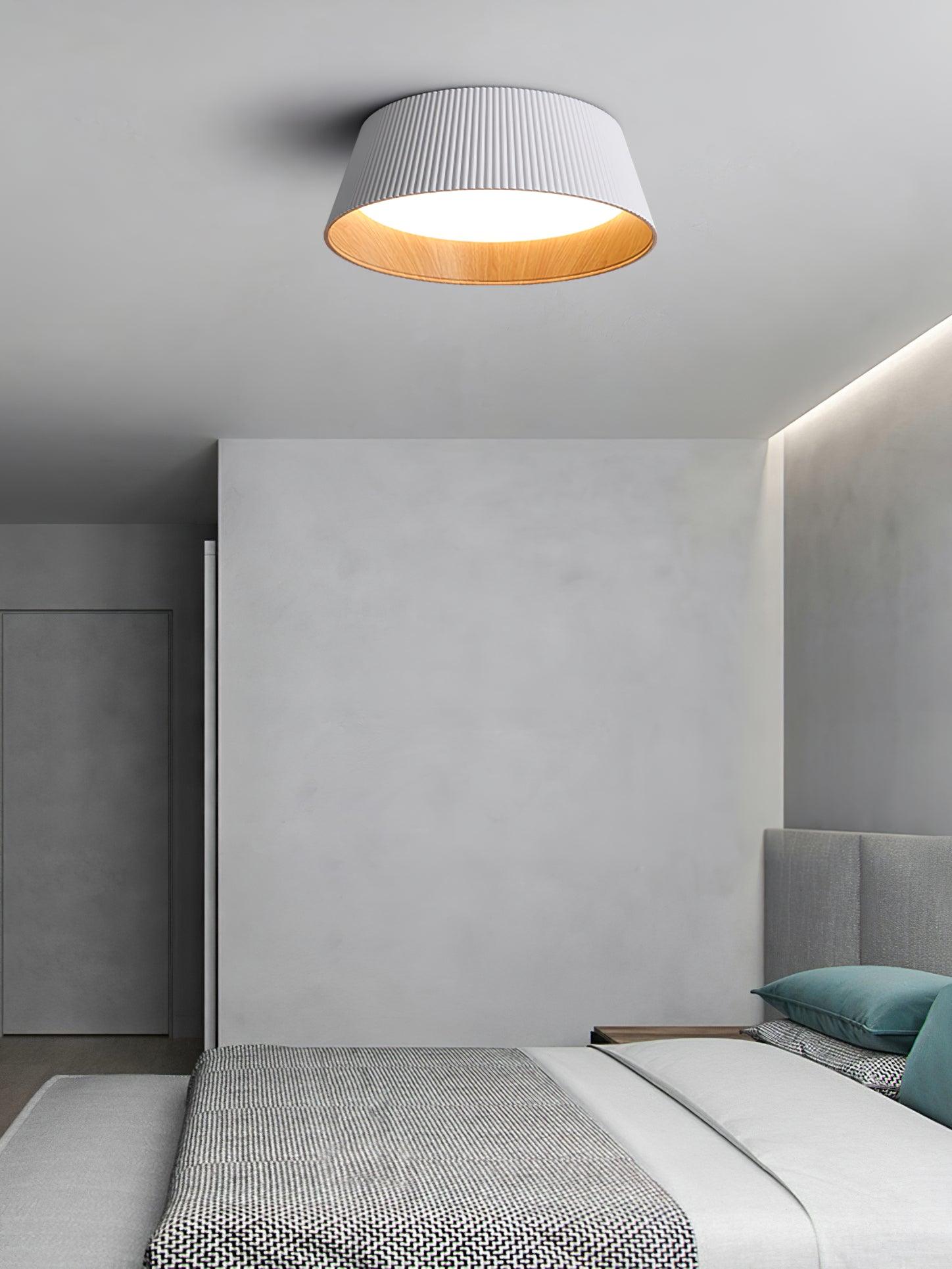 Drum-shaped flush ceiling light with a ribbed white outer, a wood-look inner rim and a white diffuser fitted to a ceiling in a bedroom with a concrete wall.