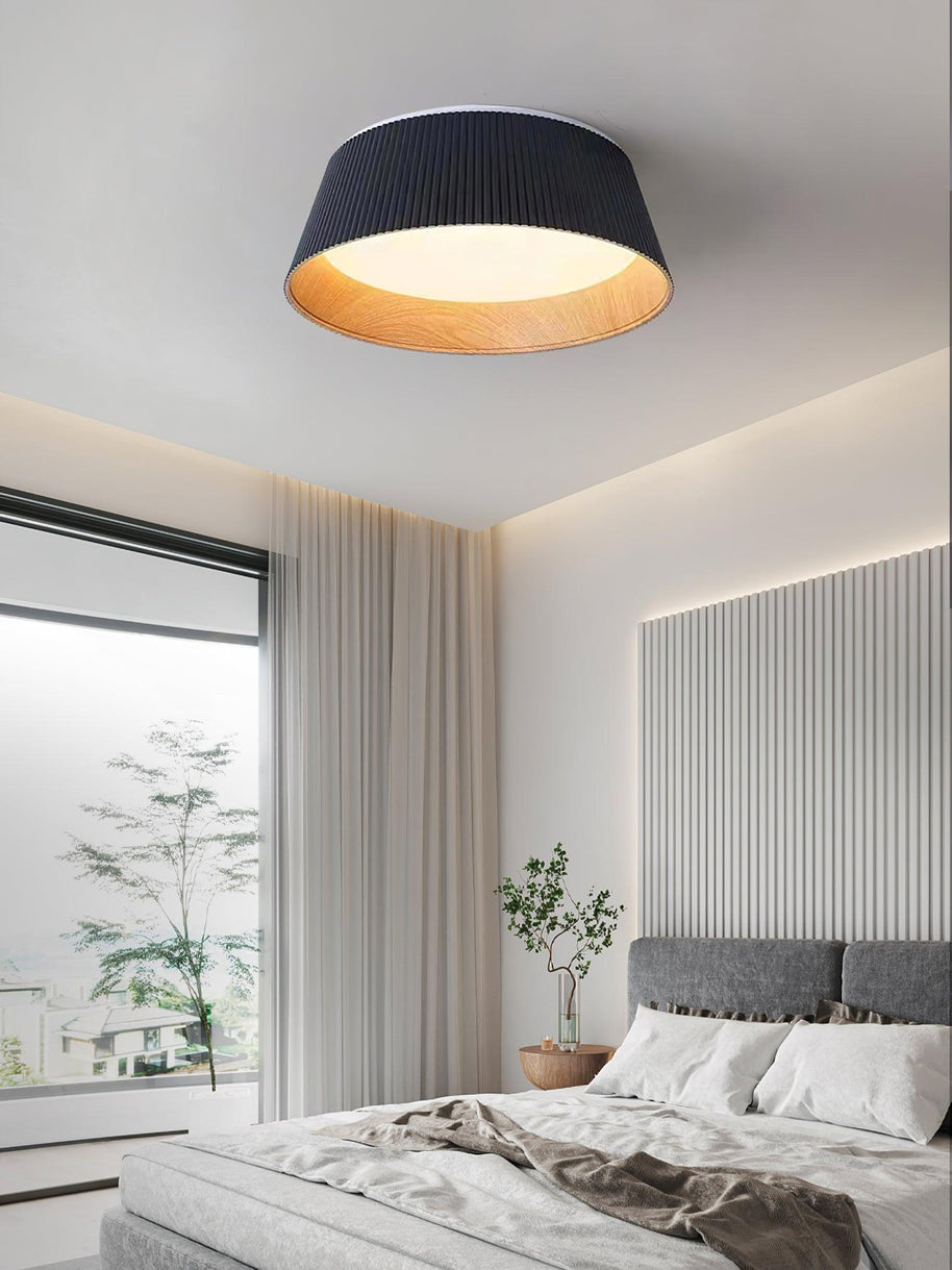 Drum-shaped flush ceiling light with a ribbed black outer, a wood-look inner rim and a white diffuser fitted above a bed by a full-height window.