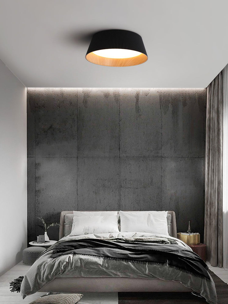 Drum-shaped flush ceiling light with a ribbed black outer, a wood-look inner rim and a white diffuser fitted above a bed against a dark textured wall.
