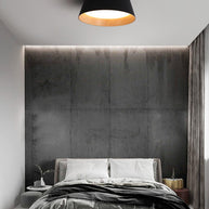 Drum-shaped flush ceiling light with a ribbed black outer, a wood-look inner rim and a white diffuser fitted above a bed against a dark textured wall.
