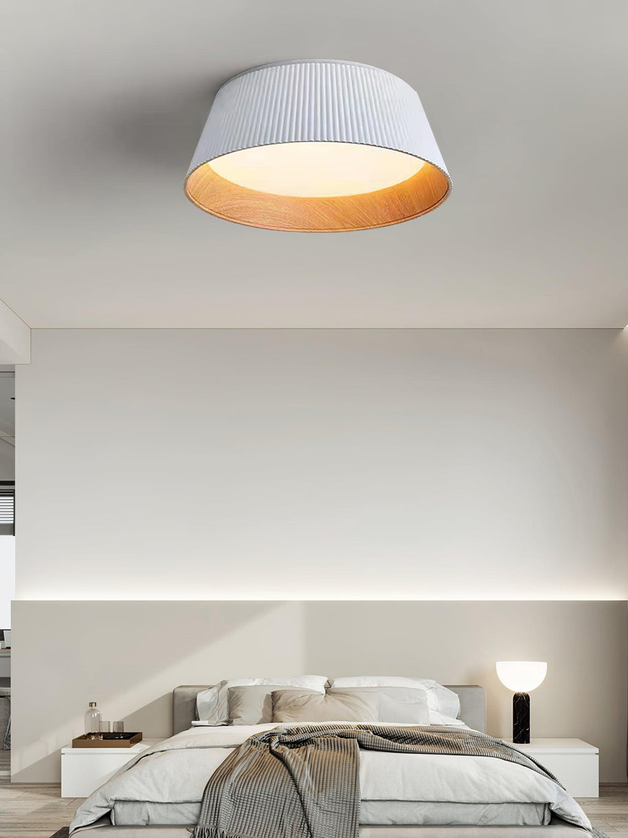 Drum-shaped flush ceiling light with a ribbed shade, a wood-look inner rim and a white diffuser, white ribbed shade, lit, fitted to a bedroom ceiling, seen from a little further back.