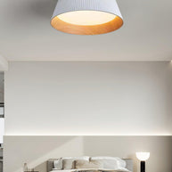 Drum-shaped flush ceiling light with a ribbed shade, a wood-look inner rim and a white diffuser, white ribbed shade, lit, fitted to a bedroom ceiling, seen from a little further back.