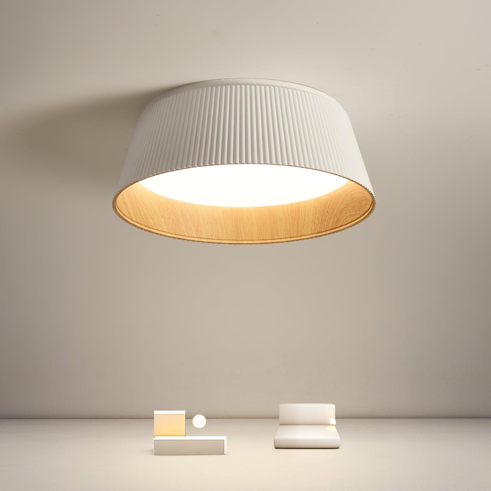 Drum-shaped flush ceiling light with a ribbed white outer, a wood-look inner rim and a white diffuser lit on a pale wall above small objects.