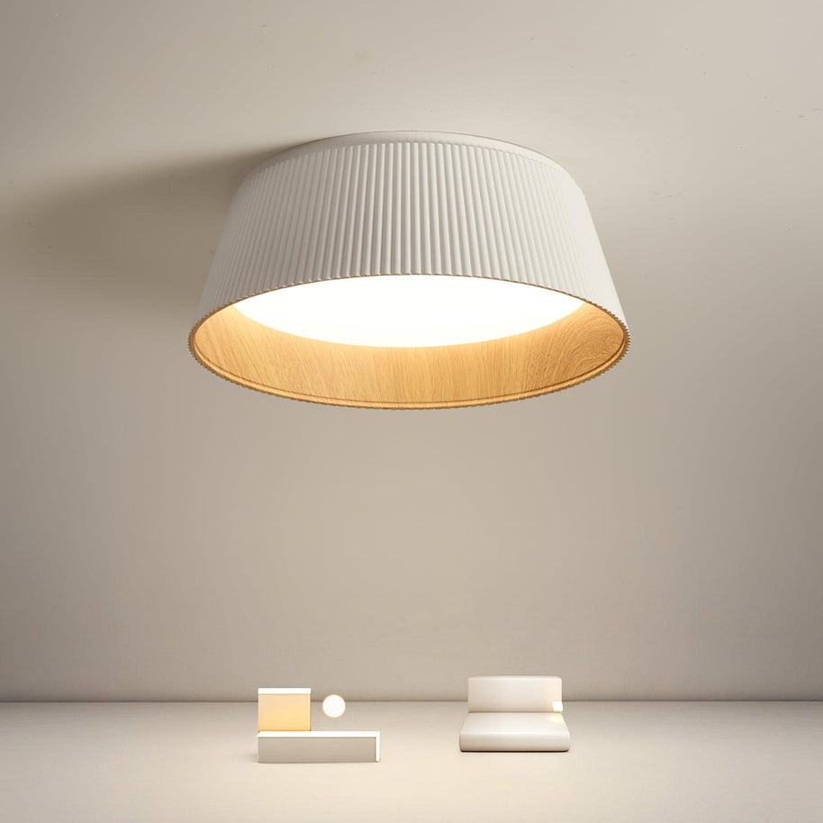Drum-shaped flush ceiling light with a ribbed white outer, a wood-look inner rim and a white diffuser lit on a pale wall above small objects.
