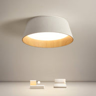 Drum-shaped flush ceiling light with a ribbed white outer, a wood-look inner rim and a white diffuser lit on a pale wall above small objects.