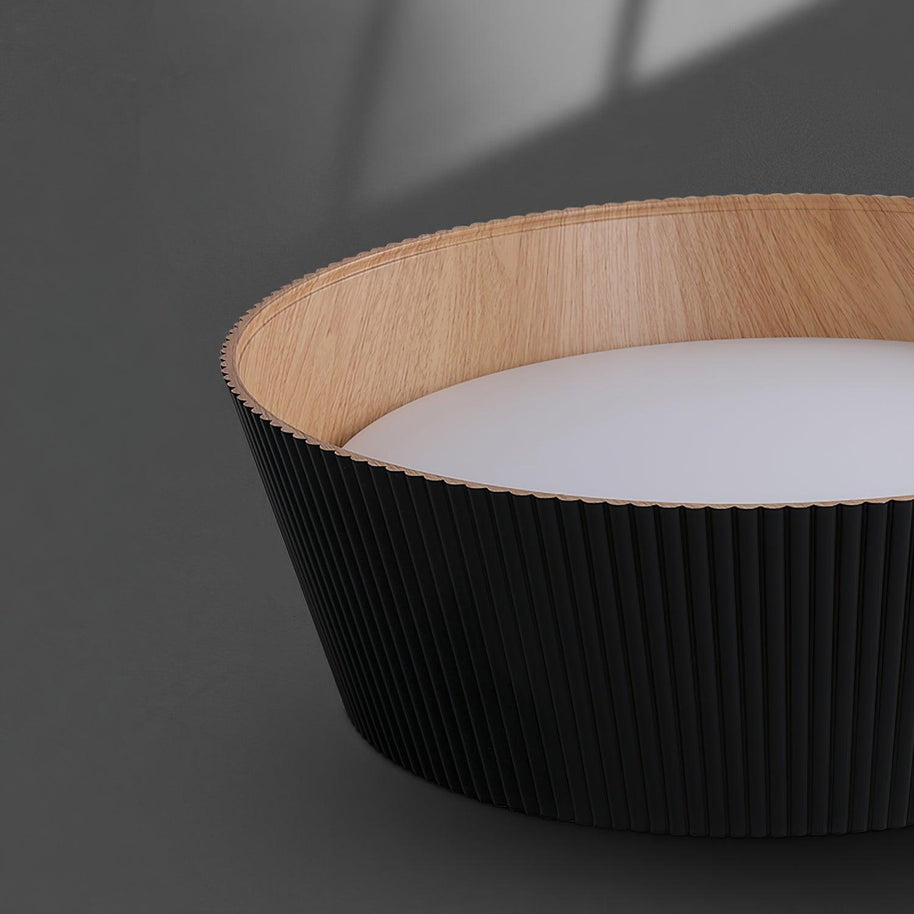 Drum-shaped flush ceiling light with a ribbed black outer, a wood-look inner rim and a white diffuser, unlit, seen from below on a dark background.