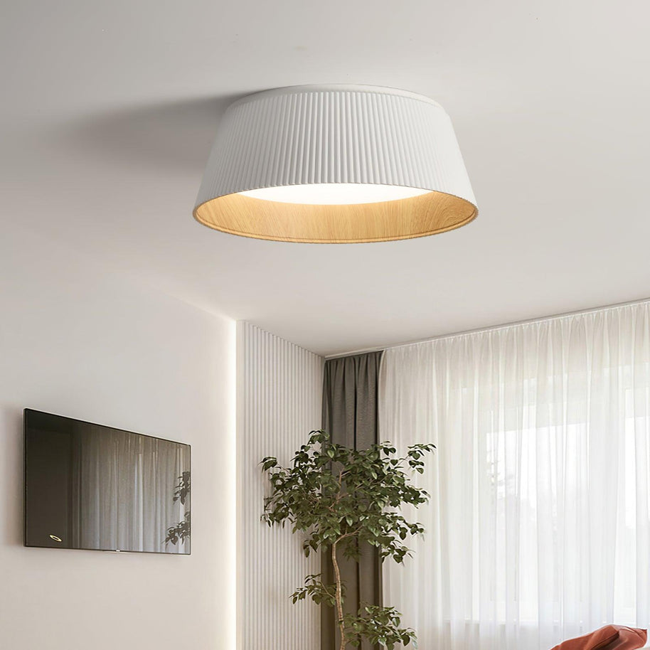 Drum-shaped flush ceiling light with a ribbed shade, a wood-look inner rim and a white diffuser, white ribbed shade, lit, fitted to a ceiling above a television and a plant.