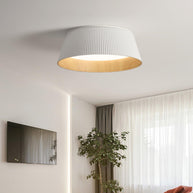 Drum-shaped flush ceiling light with a ribbed shade, a wood-look inner rim and a white diffuser, white ribbed shade, lit, fitted to a ceiling above a television and a plant.