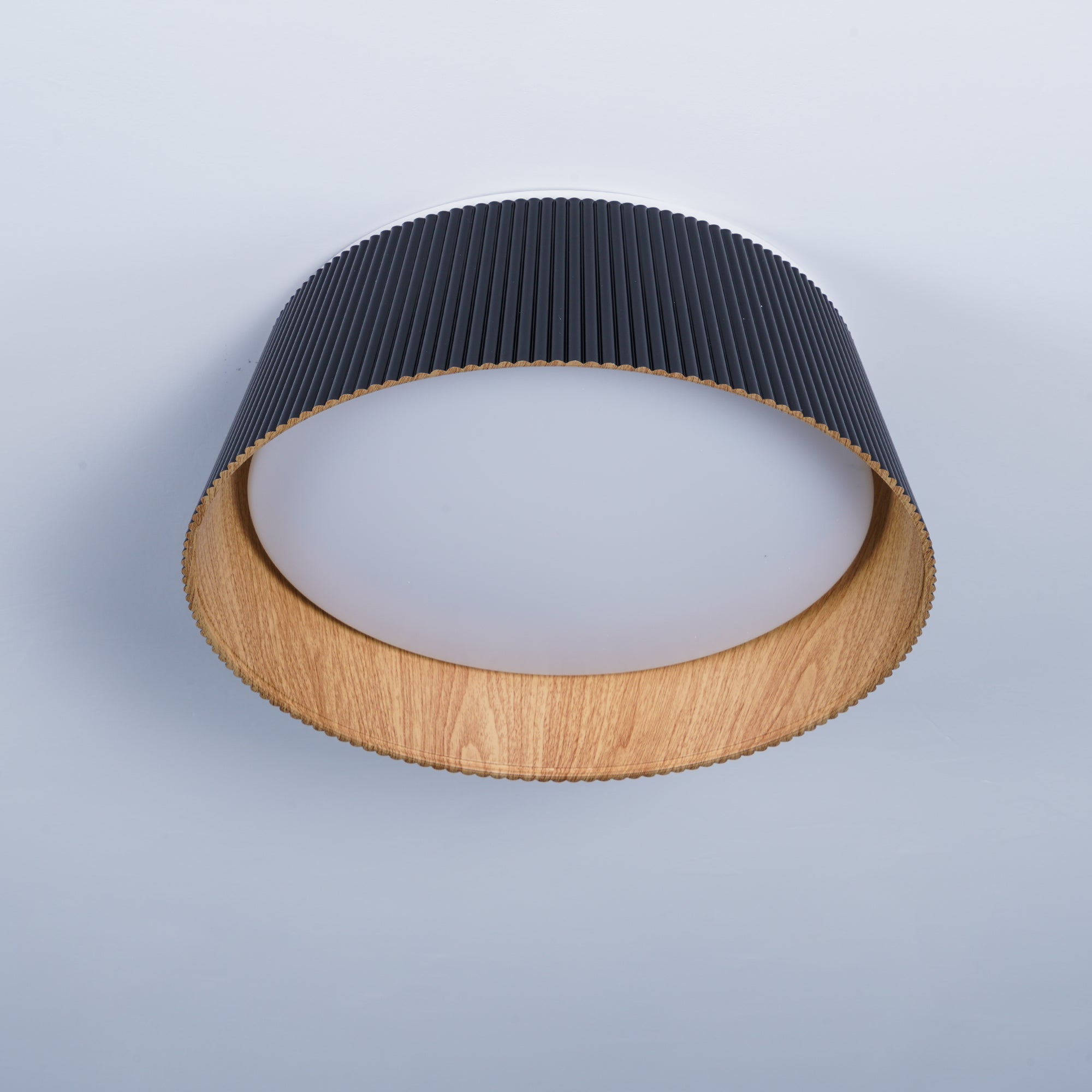 Drum-shaped flush ceiling light with a ribbed black outer, a wood-look inner rim and a white diffuser, seen from below at an angle.