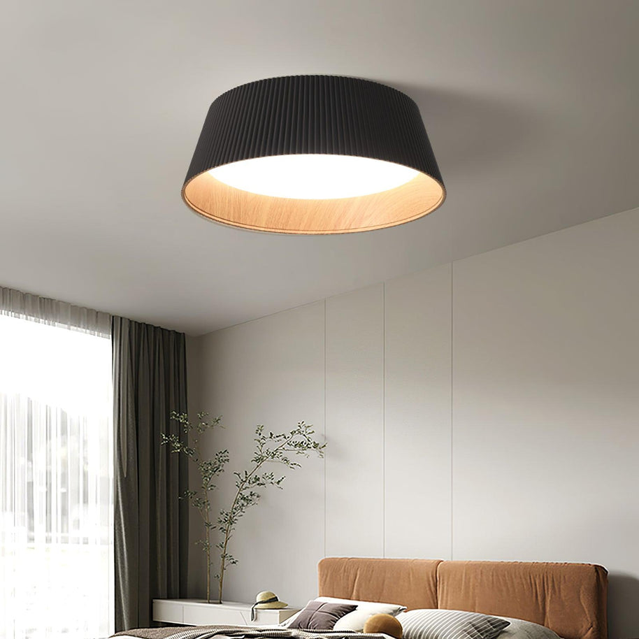 Drum-shaped flush ceiling light with a ribbed black outer, a wood-look inner rim and a white diffuser fitted above a bed beside a curtain.