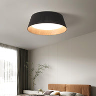Drum-shaped flush ceiling light with a ribbed black outer, a wood-look inner rim and a white diffuser fitted above a bed beside a curtain.
