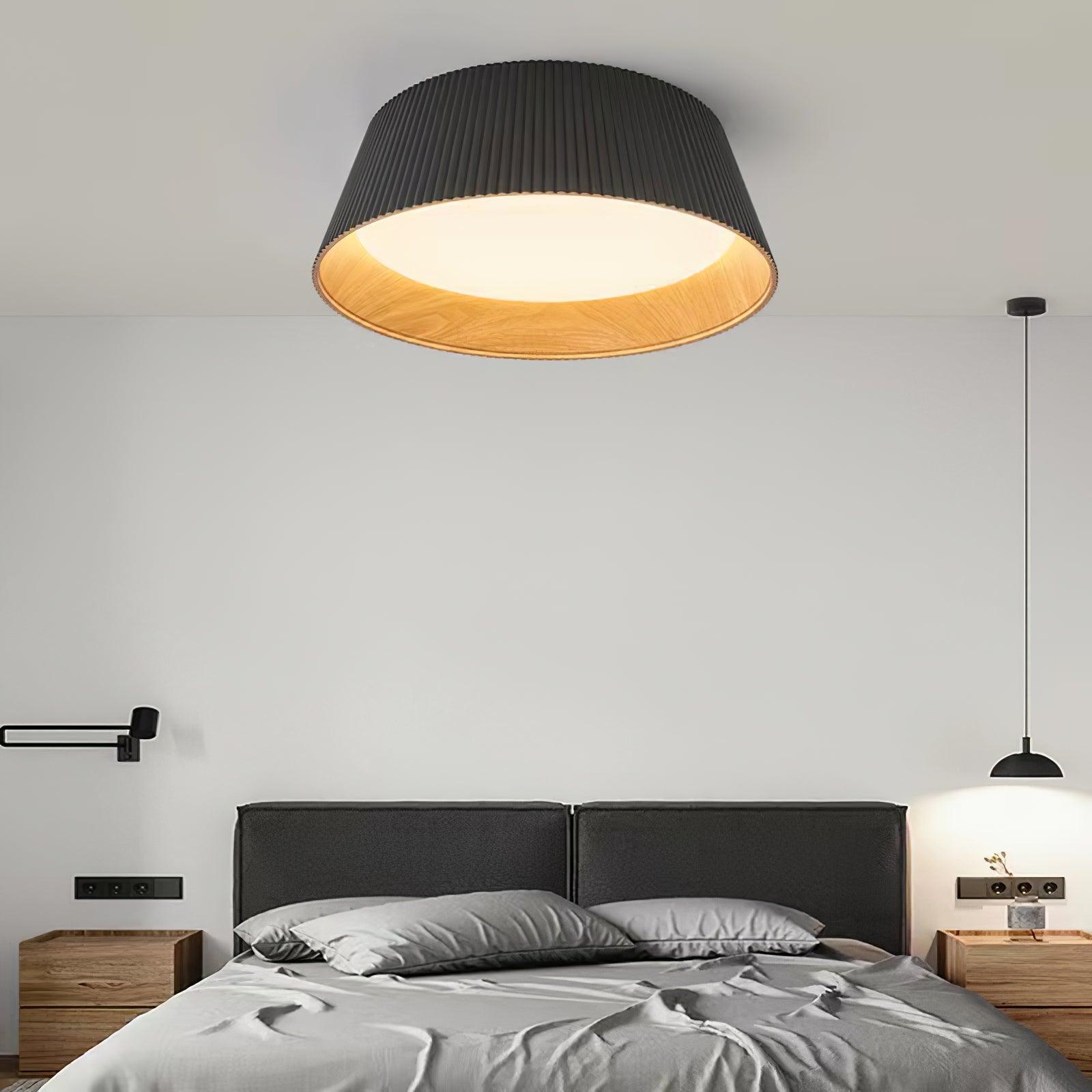 Drum-shaped flush ceiling light with a ribbed black outer, a wood-look inner rim and a white diffuser fitted above a bed in a grey bedroom.