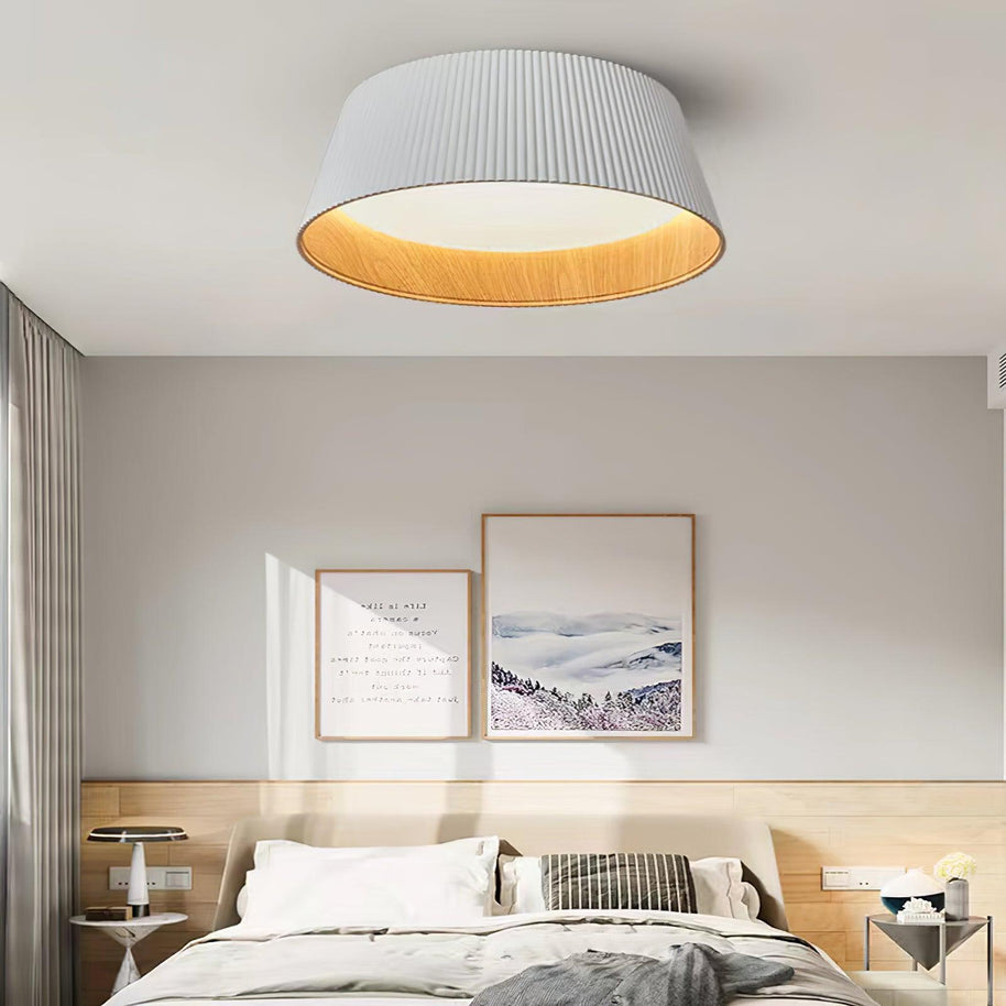 Drum-shaped flush ceiling light with a ribbed white outer, a wood-look inner rim and a white diffuser fitted above a bed with framed prints behind.