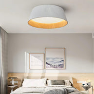Drum-shaped flush ceiling light with a ribbed white outer, a wood-look inner rim and a white diffuser fitted above a bed with framed prints behind.