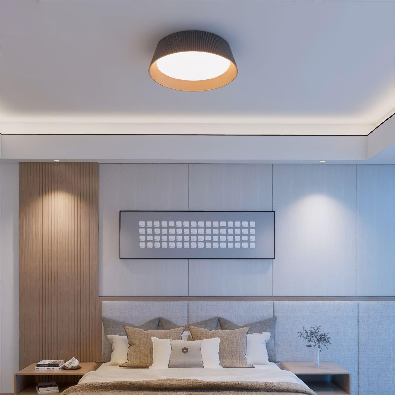 Drum-shaped flush ceiling light with a ribbed black outer, a wood-look inner rim and a white diffuser fitted to a ceiling in a bedroom.