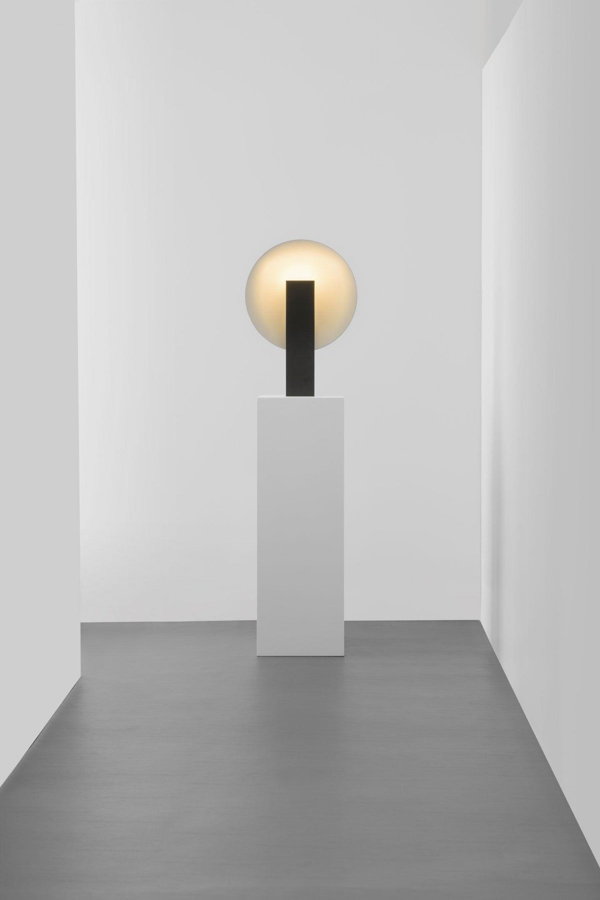 Orbelyn Table Lamp, lit, on a tall white plinth in a white gallery corridor