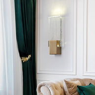 Wall light with a ribbed clear glass panel and a brushed brass mount lit on a panelled wall beside a green curtain.