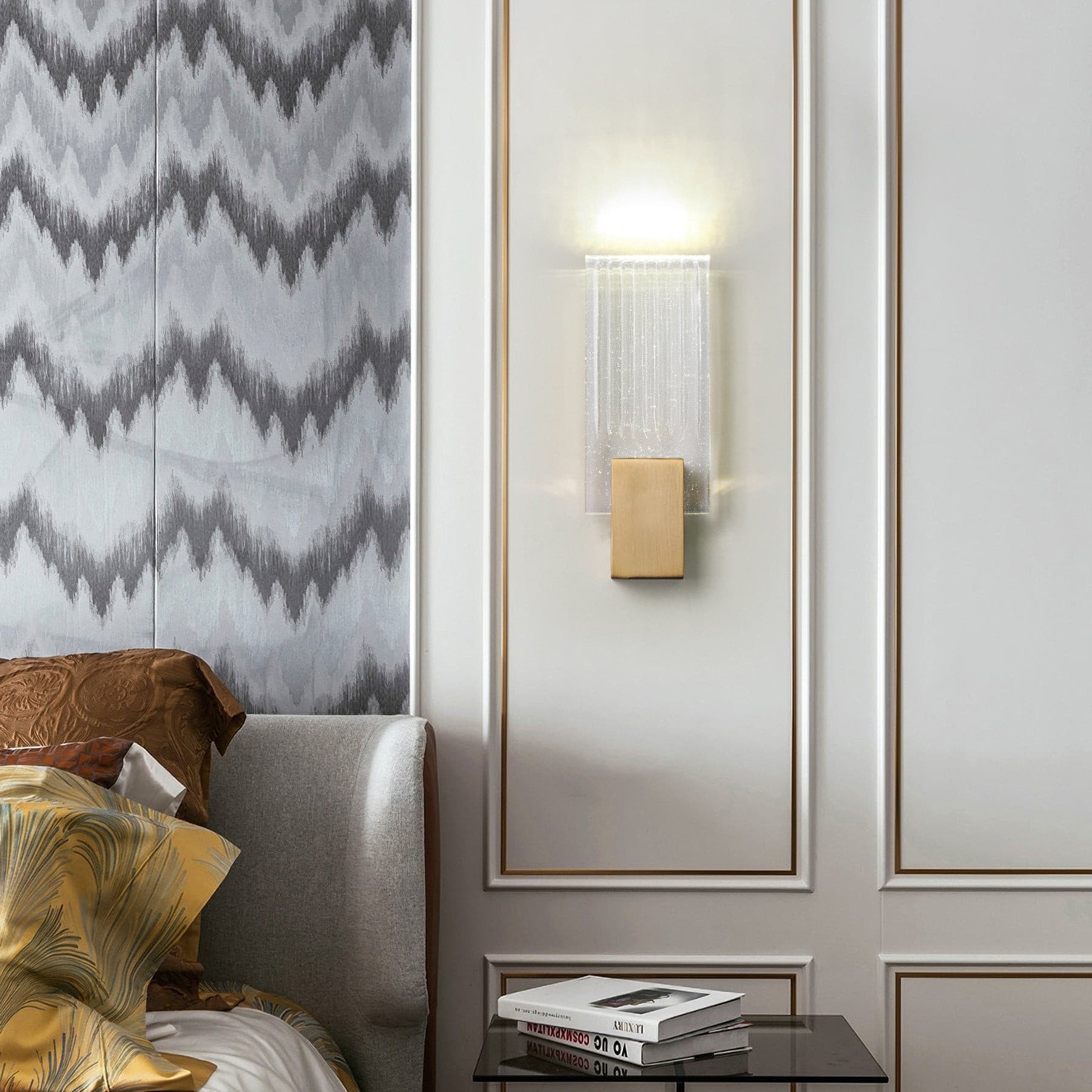 Wall light with a ribbed clear glass panel and a brushed brass mount lit on a panelled bedroom wall.