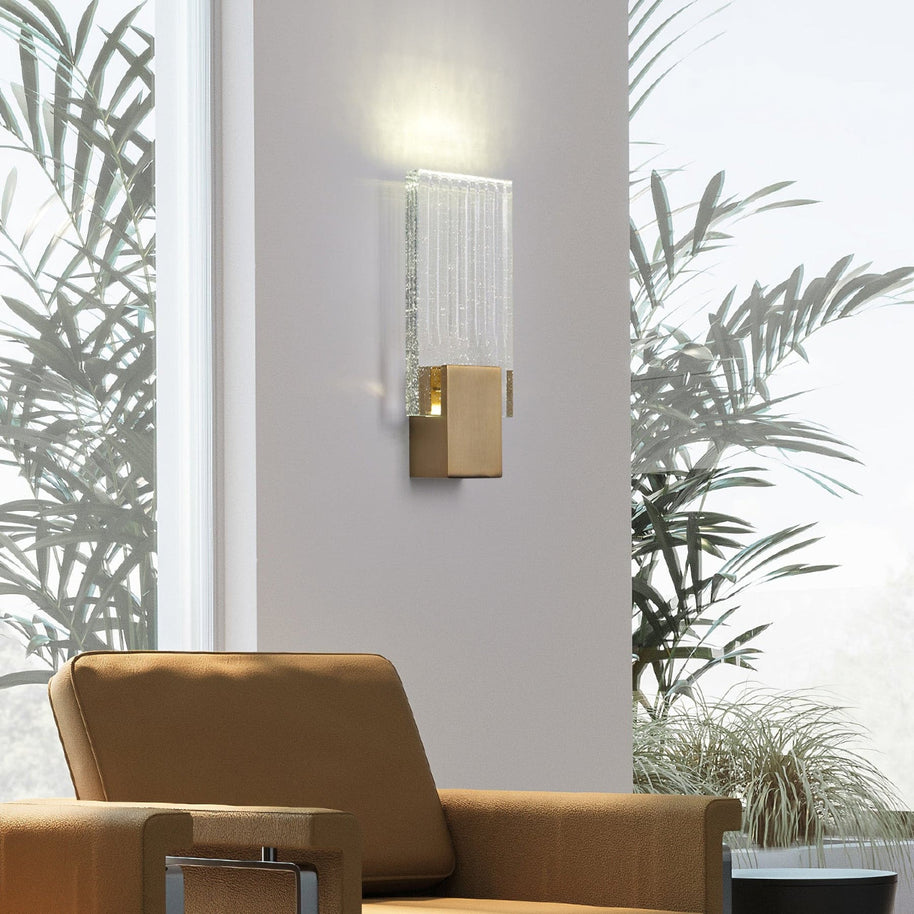 Wall light with a ribbed clear glass panel and a brushed brass mount lit on a wall beside a tan armchair and palms.