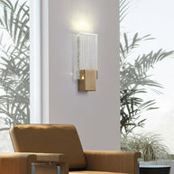 Wall light with a ribbed clear glass panel and a brushed brass mount lit on a wall beside a tan armchair and palms.