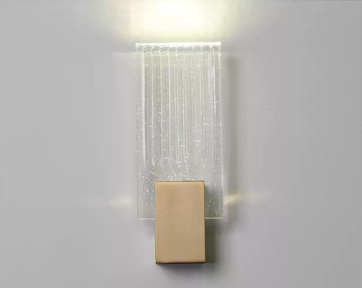 Wall light with a ribbed clear glass panel and a brushed brass mount lit, seen close against a pale wall.