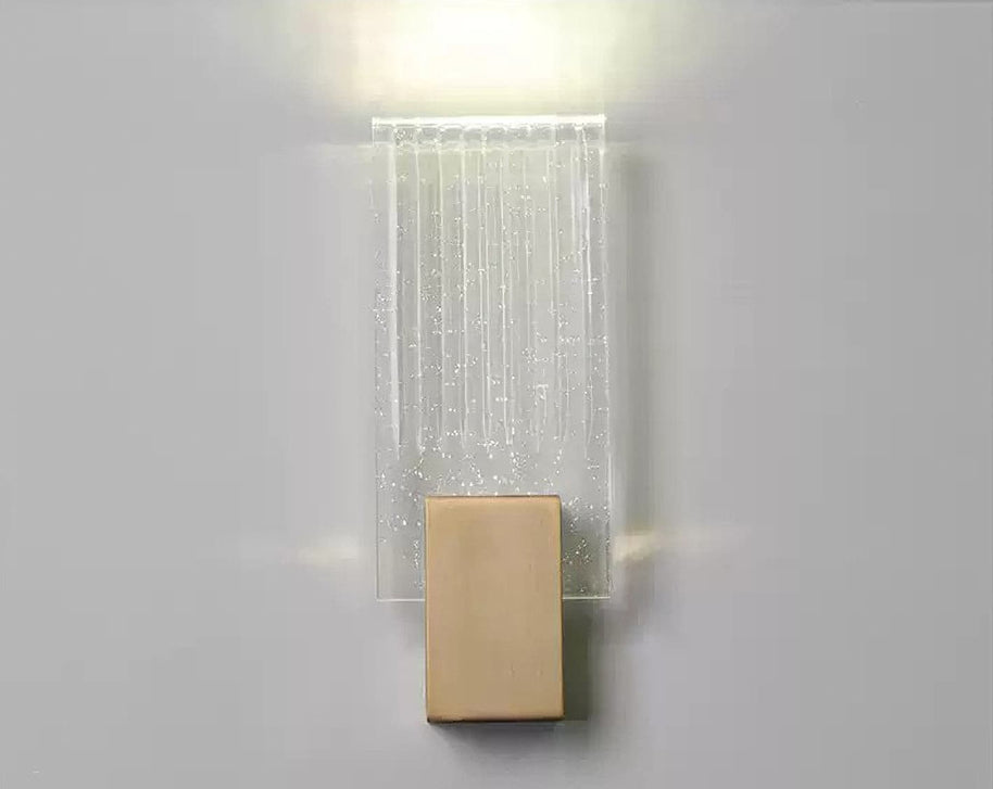 Wall light with a ribbed clear glass panel and a brushed brass mount lit, seen close against a pale wall.