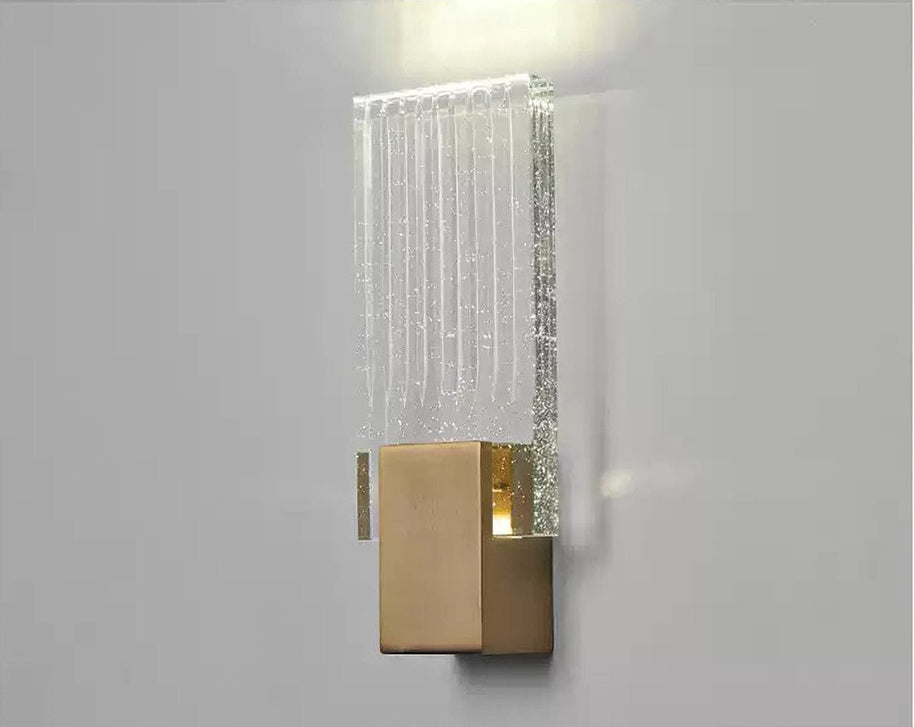 Wall light with a ribbed clear glass panel and a brushed brass mount lit against a pale wall, seen close.