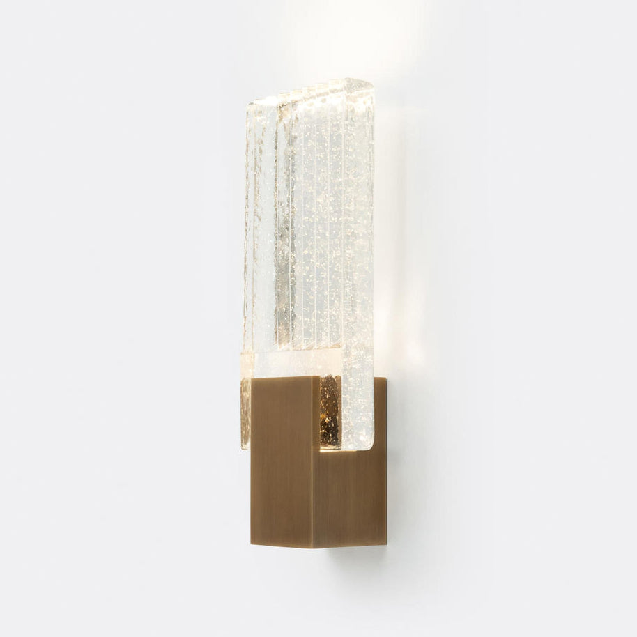 Wall light with a ribbed clear glass panel and a gold-toned metal mount, lit, on a white background, with light spilling above and below the glass.