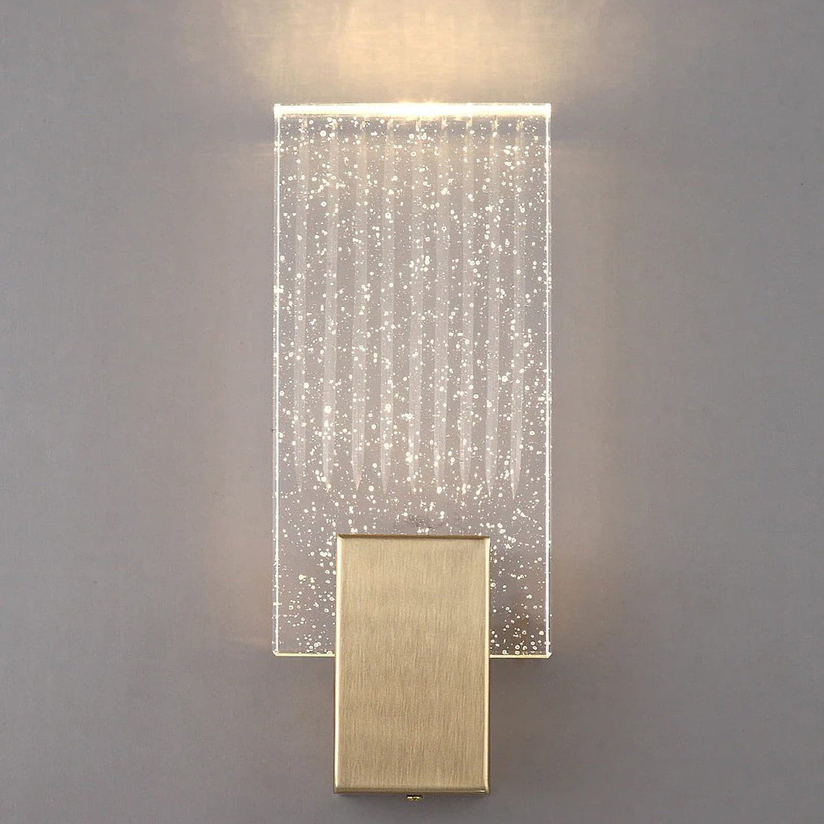 Wall light with a ribbed clear glass panel and a brushed brass mount lit, shown close on a plain wall.
